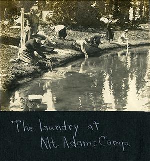 Women in skirts and sun hats kneel along a lakeshore washing clothes